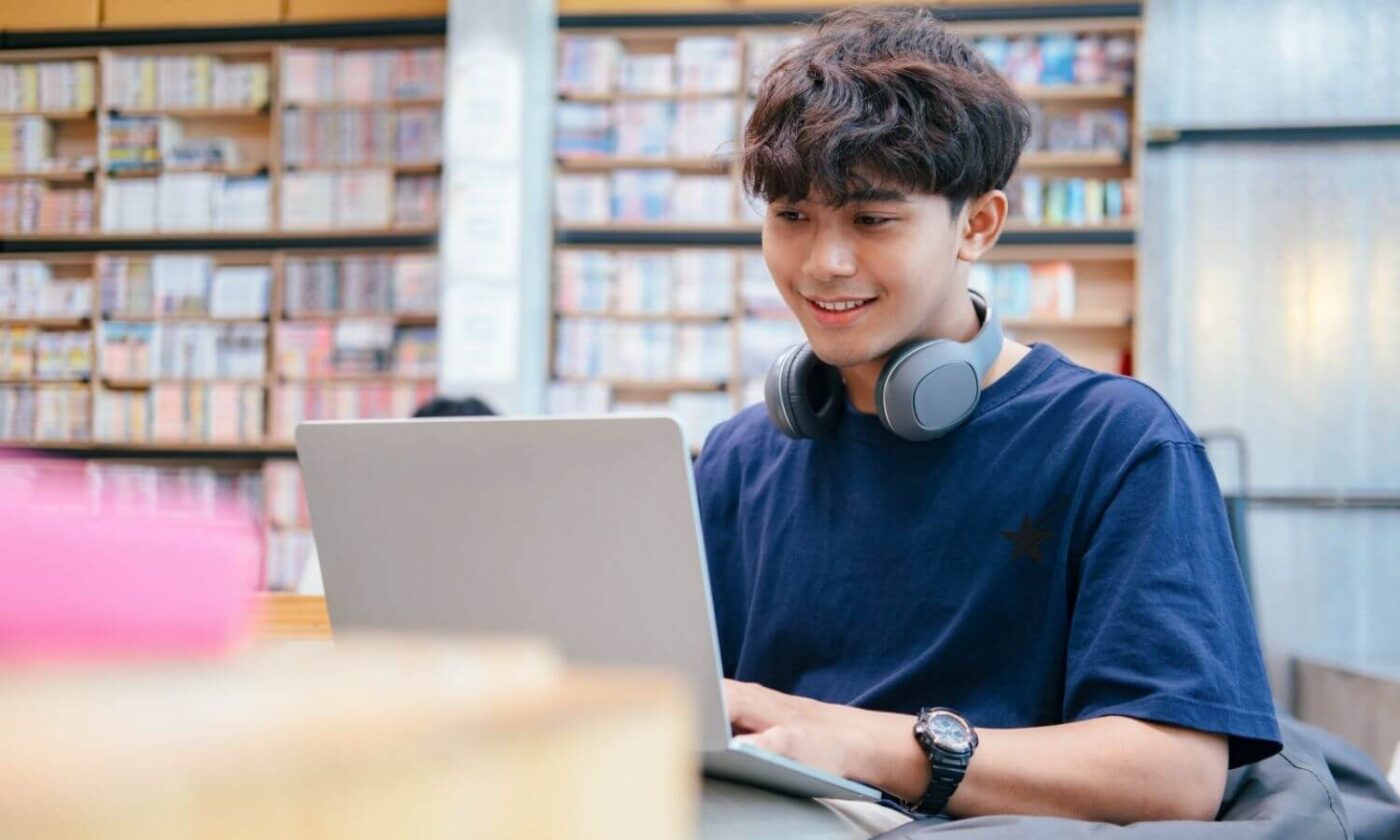Young man working on a laptop inside a modern public library, bookshelves in the background and headphones around his neck.
