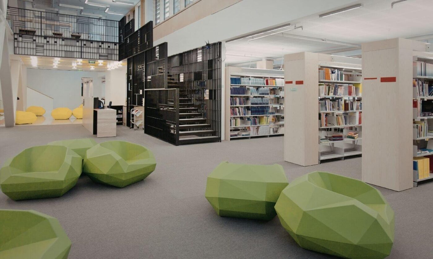 Modern public library interior with open bookshelves, black metal staircase, and geometric green seating in a bright reading area.